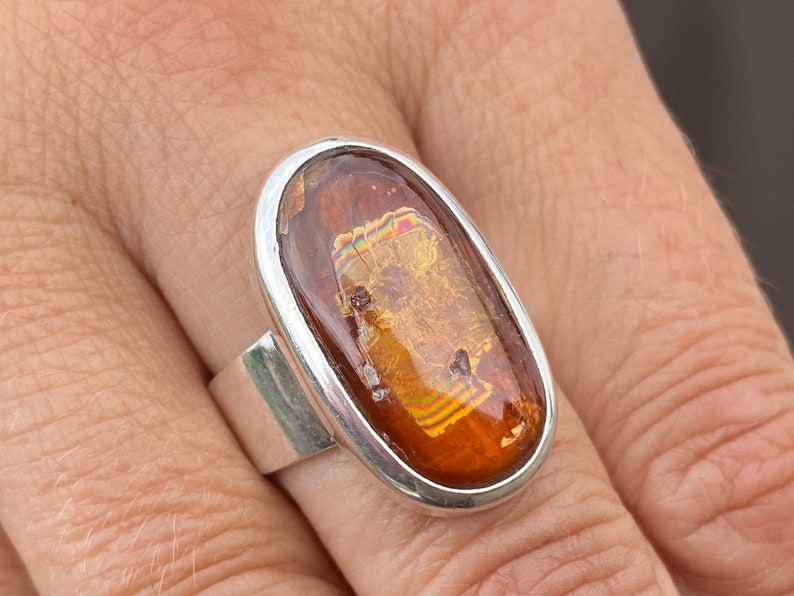 May include: A silver ring with an oval-shaped amber stone. The stone has a unique iridescent pattern and is set in a silver bezel.