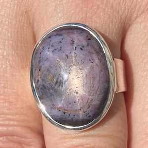 May include: A silver ring with a large, oval gemstone in shades of lavender and grey. The stone has a polished surface with dark speckles and a subtle sheen. The ring's band is a simple, polished silver.