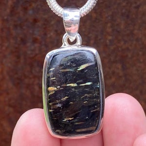 May include: A rectangular pendant with a black stone, featuring gold and green streaks, set in a silver bezel. The pendant hangs from a silver chain. The jewelry is held by a person's fingers.