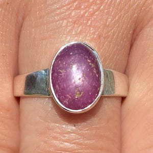 May include: A silver ring featuring an oval, purplish-pink gemstone. The gemstone has a textured surface and is set in a simple, polished silver band. The ring is shown on a finger, highlighting the jewelry's design and color.