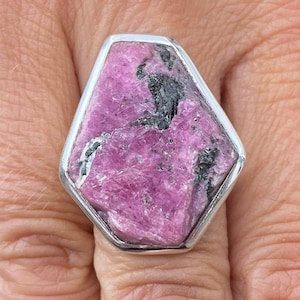 May include: A silver ring with a large, pink, faceted gemstone set in a hexagonal bezel. The gemstone has black inclusions.