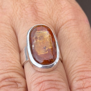 May include: A silver ring with a large oval-shaped amber stone set in a bezel setting. The stone has a golden hue and is translucent.