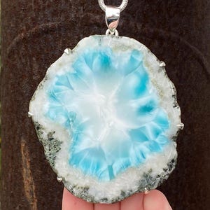 May include: A large, irregular, flat pendant featuring a central vibrant blue and white stone slice with a translucent, crystalline appearance. The stone is encased in a silver-toned metal prong setting and hangs from a silver-toned chain necklace.