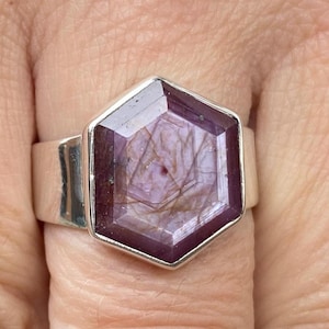 May include: A silver ring with a large, hexagonal, purplish-red gemstone. The gemstone has a distinctive internal pattern. The ring's band is wide and smooth. The ring is a piece of jewellery.
