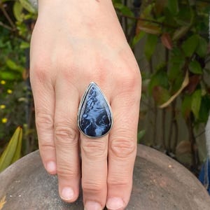May include: A silver ring with a large, teardrop-shaped gemstone. The stone features a swirling pattern of blue and white, set in a silver band. The ring is displayed on a hand.