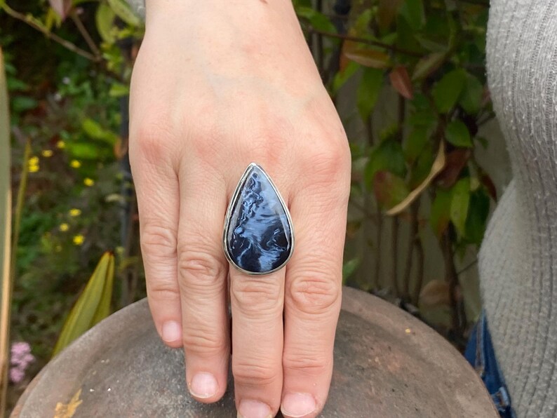 May include: A silver ring with a large, teardrop-shaped gemstone. The stone features a swirling pattern of blue and white, set in a silver band. The ring is displayed on a hand.
