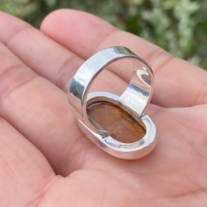 May include: A silver ring with a large oval-shaped tiger's eye gemstone. The ring is set in a bezel setting and has a polished finish.
