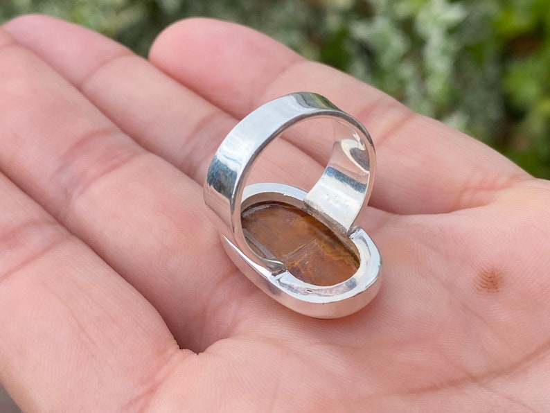 May include: A silver ring with a large oval-shaped tiger's eye gemstone. The ring is set in a bezel setting and has a polished finish.