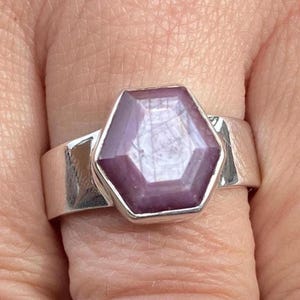 May include: A silver ring with a large, hexagonal, purplish-pink gemstone. The gemstone is set in a wide, polished silver band. The ring's design is simple yet elegant, highlighting the unique shape and colour of the stone.