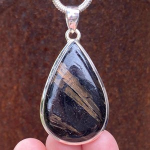 May include: A teardrop-shaped pendant with a silver setting and chain. The pendant features a polished black stone with brown inclusions. The jewelry is displayed against a blurred, brown background.