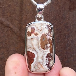 May include: A rectangular pendant with a silver frame and chain. The pendant features a stone with a pattern of white, brown, and red hues. The stone is set in a polished silver bezel.