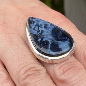 May include: A large, teardrop-shaped ring featuring a polished blue gemstone with white marbling set in a silver band. The ring is displayed on a person's finger, showcasing the stone's unique pattern and the ring's design.