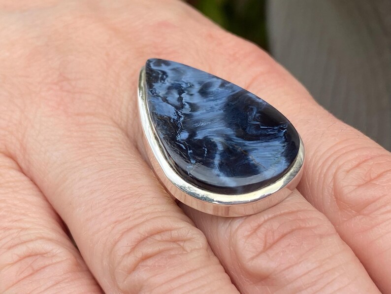 May include: A large, teardrop-shaped ring featuring a polished blue gemstone with white marbling set in a silver band. The ring is displayed on a person's finger, showcasing the stone's unique pattern and the ring's design.