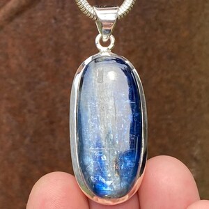 May include: An oval-shaped blue kyanite gemstone pendant set in a polished silver bezel. The stone features deep blue and translucent white streaks throughout its surface. The pendant is attached to a silver snake chain necklace via a silver bail.