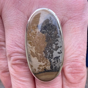 May include: Oval-shaped ring featuring a large, polished stone with a unique pattern of brown, beige, and gray hues. The stone is set in a silver-colored metal band. The ring's design showcases natural stone jewelry.