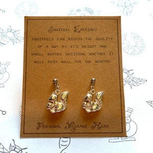 Könnte beinhalten: Ein Paar goldfarbene Eichhörnchen-Ohrringe. Die Eichhörnchen halten Nüsse in ihren Pfoten. Die Ohrringe befinden sich auf einer braunen Karte mit dem Text "SQUIRREL EARRINGS" und "PERSONAL MESSAGE HERE".