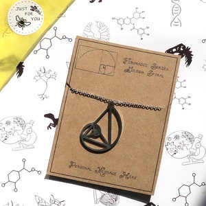 以下が含まれることがあります： 「フィボナッチ数列 黄金螺旋」と書かれた茶色のカードと、黄金螺旋の形をした黒い木製ペンダント。ペンダントは銀色のチェーンに付いています。カードの下部には「パーソナルメッセージはこちら」と書いてあります。