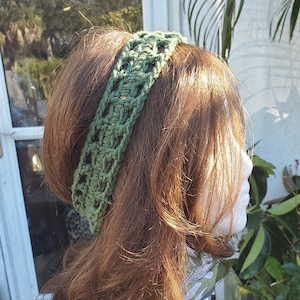 May include: A green crocheted headband with an open weave design is displayed on a mannequin head with auburn hair. The headband is a solid color and appears to be handmade. The background includes plants and outdoor elements.