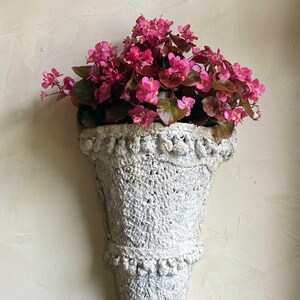 May include: A white, textured wall planter shaped like a cornucopia, filled with vibrant pink flowers and green foliage. The planter has decorative trim and is mounted on a light-colored wall. A decorative piece for the home.