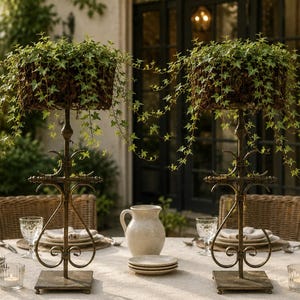 May include: Two tall, bronze-colored metal stands with square bases and ornate scrollwork, each supporting a rectangular basket filled with trailing green ivy plants. The stands are positioned on a table set with plates and glassware.
