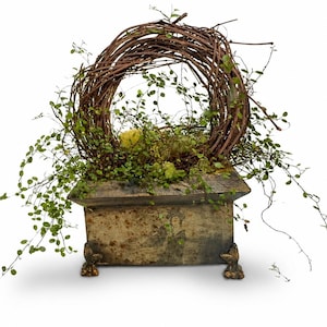 May include: A rustic, square planter with a weathered appearance, adorned with a circular twig wreath and cascading green foliage. The planter rests on decorative feet, creating a natural, earthy aesthetic.