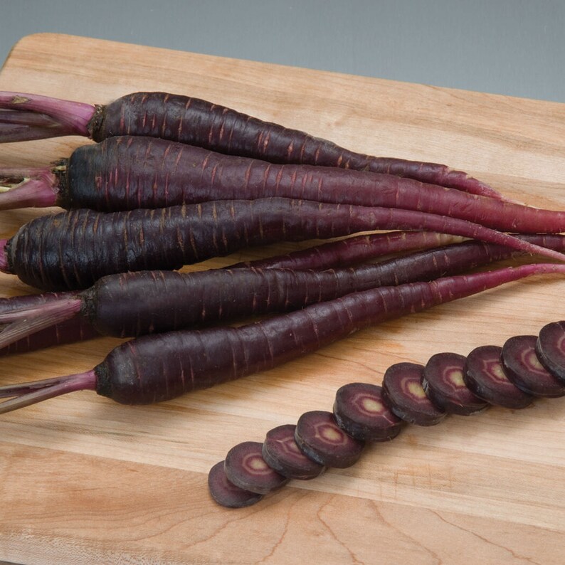 May include: A bunch of purple carrots on a wooden cutting board. Some of the carrots are sliced and show the purple flesh.