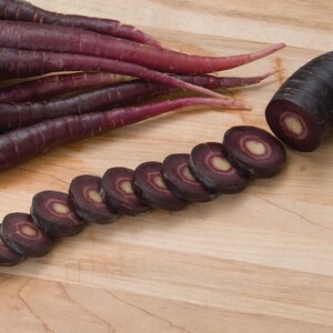 May include: Purple carrots sliced on a wooden cutting board. The carrots are arranged in a line with the cut ends facing the viewer. The carrots are a deep purple color with a white core.