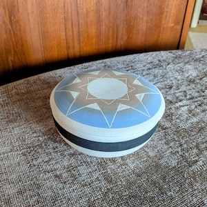 May include: A round ceramic box with a blue and white starburst design. The box has a black band around the edge.
