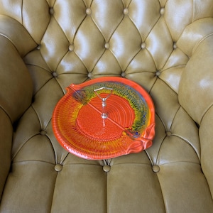 May include: A vibrant orange ceramic serving dish with a silver handle. The dish features a textured surface and a colorful rim with yellow, blue, and green accents. The dish is placed on a tufted, tan leather chair.