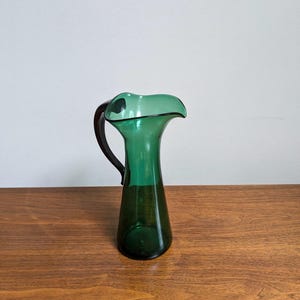 May include: A green glass pitcher with a dark handle. The pitcher has a flared rim and a dark green color. It is sitting on a wooden surface.