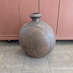 May include: A large, round, antique metal vessel with a short, cylindrical neck. The vessel has a dark, aged patina with visible seams and a slightly flattened base. The overall shape is reminiscent of a large jug or urn.