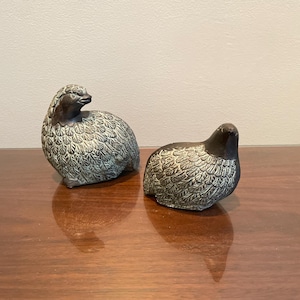 May include: Two decorative bird figurines. The birds are in a resting position, with detailed feather patterns. The figurines are primarily gray and brown, with a textured appearance. They are placed on a wooden surface.