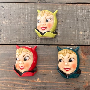 May include: Three ceramic wall decor pieces shaped like faces with devil horns. The faces are painted with a light pink complexion, large eyes, and a red smile. Each face is wearing a different colored hood: yellow, red, and blue.