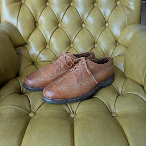 May include: A pair of light brown leather dress shoes with intricate brogue detailing and matching laces. The shoes are placed on a yellow tufted leather chair, showcasing their classic design and craftsmanship.