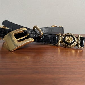 May include: Black leather belt with gold-tone buckle and lion head medallion. The belt features a textured, crocodile-like pattern. The buckle is a large, rectangular shape. The lion head medallion is round and detailed.