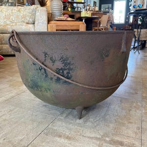 May include: A large, antique cast iron cauldron with a rounded body and three small legs. The cauldron has a rusty brown color with patches of green and a metal handle for carrying. It is likely used for cooking or decorative purposes.