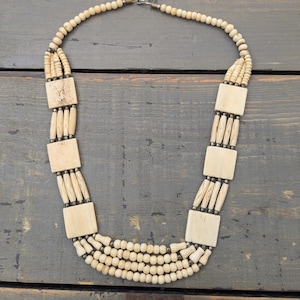 Op de afbeelding: Crèmekleurige ketting met een meerstrengs ontwerp met kleine ronde kralen en grotere vierkante en cilindrische kralen. De ketting heeft een sluiting en wordt weergegeven op een donker houten oppervlak.