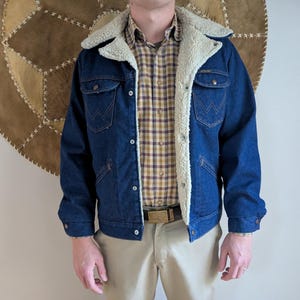 May include: A dark blue denim jacket with a cream-colored sherpa lining and collar. The jacket has two chest pockets and two side pockets. The person is wearing a plaid shirt and khaki pants. A gold belt is visible.