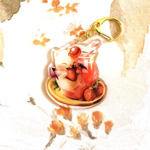 May include: A clear acrylic keychain featuring a cartoon character enjoying a strawberry drink. The keychain has a gold-colored clasp. The drink is in a glass jar with a straw, and there are fresh strawberries on the plate.