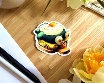 May include: A colorful sticker featuring a cartoon character designed as a burger. The burger has a dark blue and cream-colored top, with a fried egg on top. The burger sits on a wooden bun with cheese and lettuce.