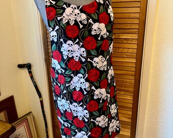 May include: A sleeveless dress with a black background, featuring a pattern of white skulls, red roses, and thorny vines. The dress is displayed on a mannequin, showcasing its design and shape.