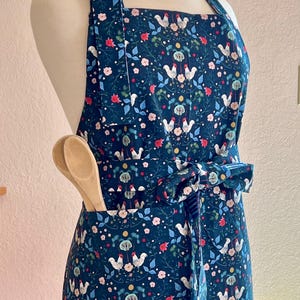 May include: A blue apron with a white rooster and floral pattern. The apron has a large pocket on the front and a tie at the waist.