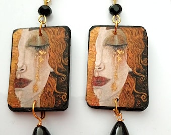 May include: A pair of gold and black earrings featuring a rectangular image of a woman with golden tears. The earrings are accented with black beads and gold triangle charms.