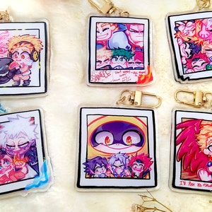 May include: Six colourful anime character photo keychain charms with gold keychains. The charms feature different characters from the anime series My Hero Academia. The characters are drawn in a chibi style and are shown in various poses. The charms are made of clear acrylic and have a glossy finish.