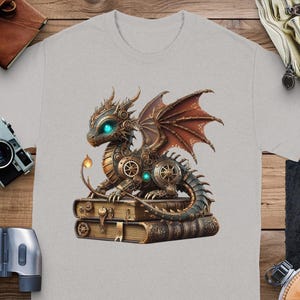 Könnte beinhalten: Ein graues T-Shirt mit einem Steampunk-Drachen-Design. Der Drache besteht aus Metallgetrieben und hat leuchtend blaue Augen. Er sitzt auf einem Stapel alter Bücher.
