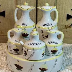 May include: A white ceramic condiment set with yellow accents. The set includes containers labeled "OIL", "VINEGAR", "SALT", "MUSTARD", and "PEPPER". Each piece features a floral design and is arranged on a matching tray.