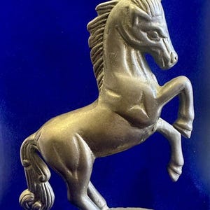 May include: A silver-colored unicorn statue in a rearing pose. The mythical creature has a detailed mane, tail, and a spiraled horn. The statue is mounted on a round base with a textured edge, set against a deep blue background.