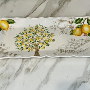 May include: Rectangular serving platter with a scalloped edge. The white ceramic platter features a lemon tree illustration, lemons, and floral accents. A wooden crate with the word "LEMON" is also depicted.