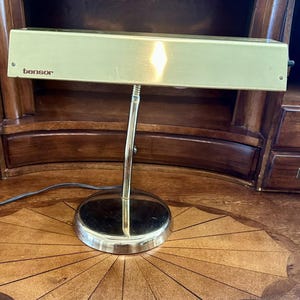 May include: A vintage Tensor desk lamp with a rectangular, light green shade and a chrome-colored, adjustable arm. The lamp has a round base and the brand name "tensor" is printed on the shade. The lamp is on a wooden desk.