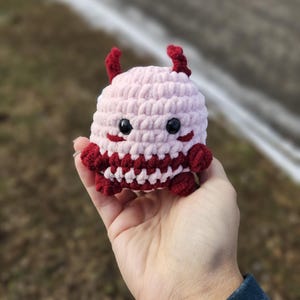 May include: A small pink and red crocheted plush toy with horns and a smiling face.